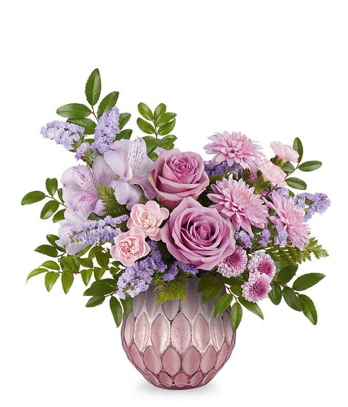Lavender roses, lavender alstroemeria, miniature pink carnations, lavender button spray chrysanthemums, and lavender sinuata statice accented with greenery in a textured pink vase.