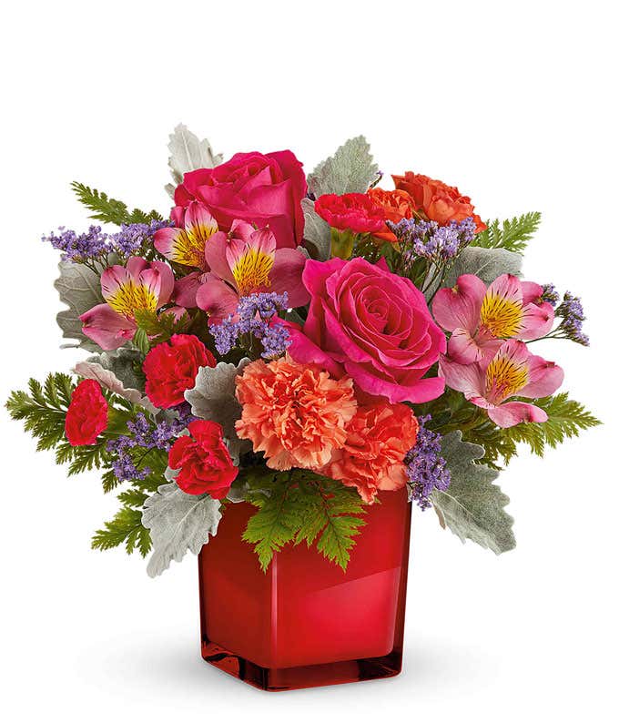 Floral arrangement with hot pink roses, pink alstroemeria, orange carnations, miniature red carnations, purple limonium, and assorted greenery in a red cube vase.
