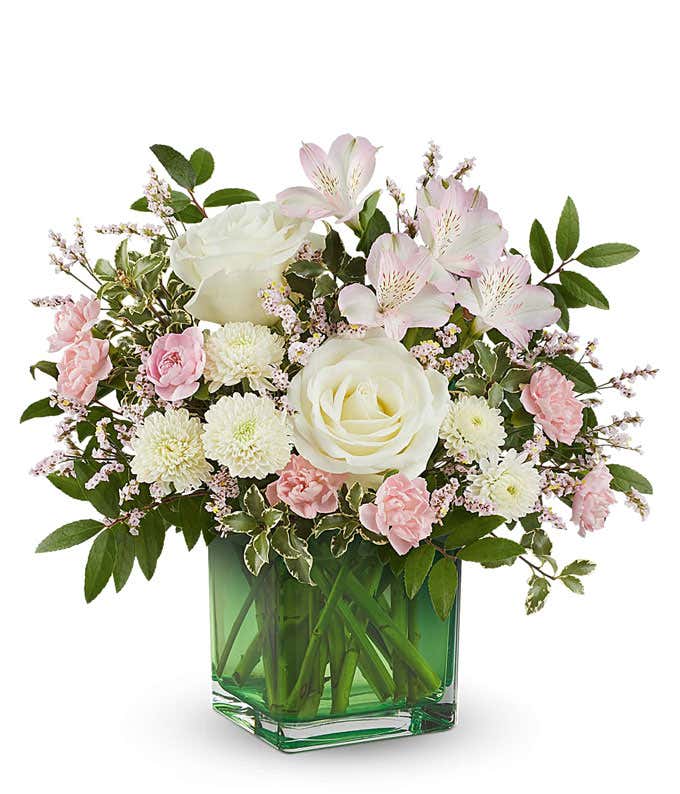 Soft pastel floral arrangement featuring crme roses, pink spray roses, pink alstroemeria, miniature pink carnations, white button spray mums, delicate filler flowers, and lush greenery in a green glass cube vase.