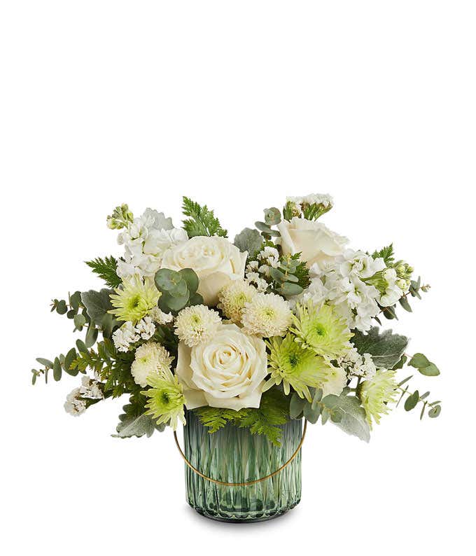 White roses, white stock, green button spray chrysanthemums, white cushion spray chrysanthemums, and assorted greenery arranged in a ribbed green glass vase