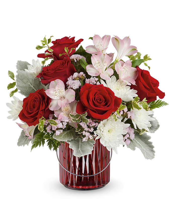 A Valentine's Day bouquet featuring red roses, light pink alstroemeria, white poms, soft pink filler flowers, dusty miller, and lush greenery, arranged in a red glass lantern with a decorative handle.