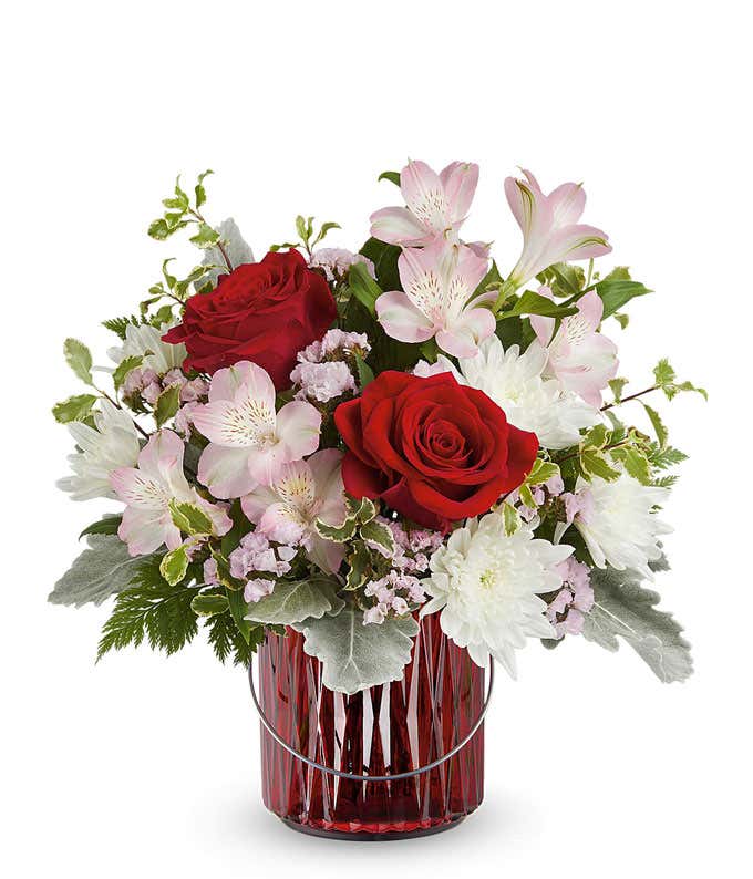 A Valentine's Day bouquet featuring red roses, light pink alstroemeria, white poms, soft pink filler flowers, dusty miller, and lush greenery, arranged in a red glass lantern with a decorative handle.