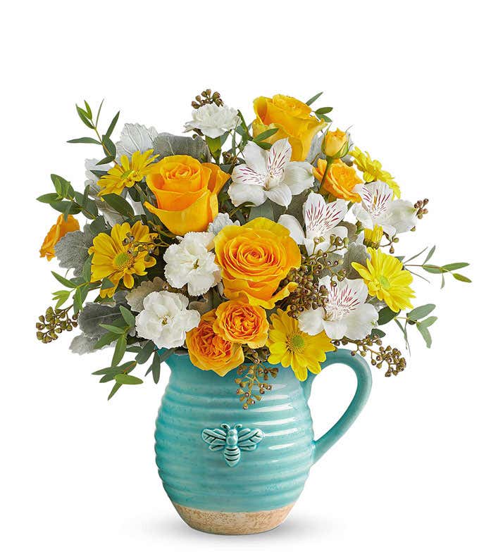 Yellow roses, white alstroemeria, miniature white carnations, yellow daisy poms, and greenery arranged in a teal ceramic pitcher with a bee design.