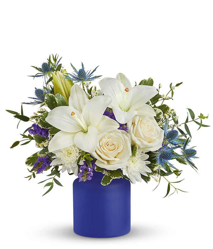 White lilies, white roses, white cushion spray chrysanthemums, blue eryngium, purple accent flowers, and greenery arranged in a matte cobalt blue cylinder vase.