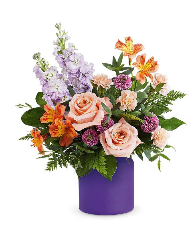 Floral arrangement with peach roses, orange alstroemeria, light lavender stock, lavender button poms, peach miniature carnations, and assorted greenery in a purple cylinder vase.
