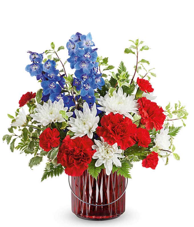 Red, white, and blue floral arrangement with red roses, white chrysanthemums, red carnations, blue delphinium, and assorted greenery in a red glass vase.