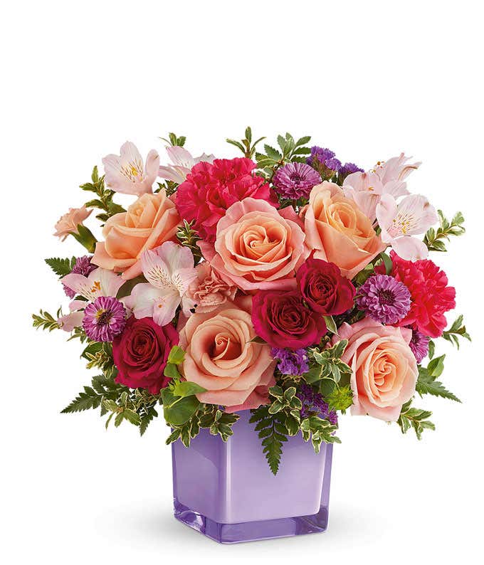 Peach Roses, hot pink spray roses, light pink alstroemeria, hot pink carnations, mini peach carnations, purple button poms, and floral greenery in a purple cube vase