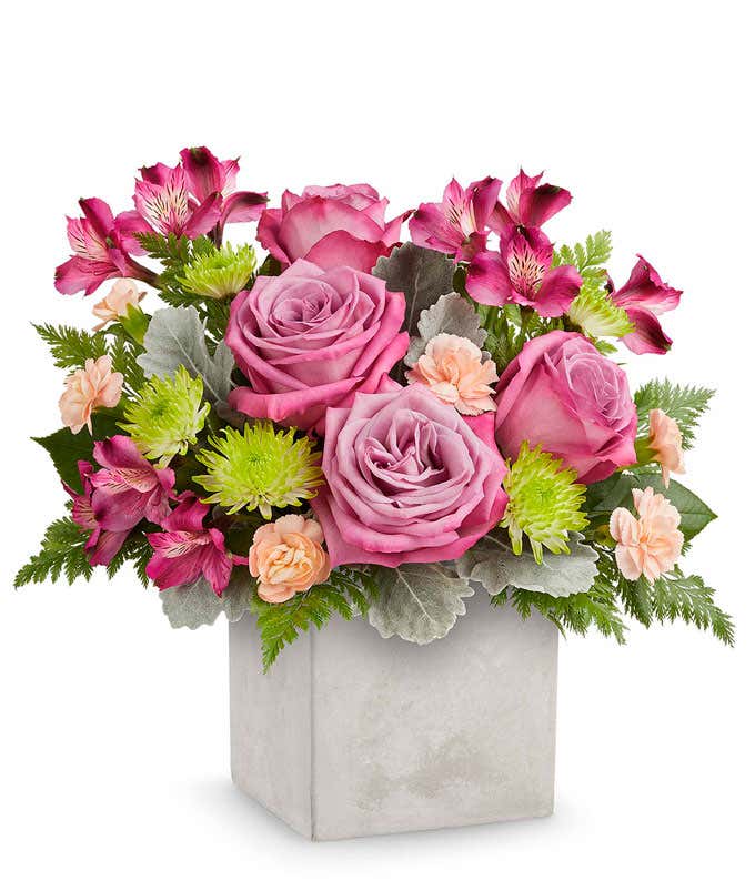 Floral arrangement with lavender roses, purple alstroemeria, mini peach carnations, green button poms, and floral greenery in a grey faux concrete look cube