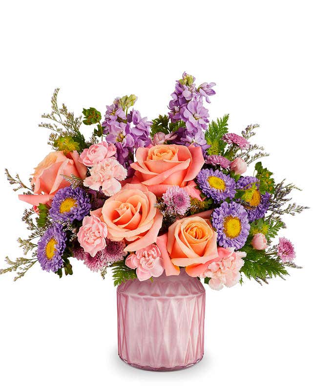 Peach roses, Mini pink carnations, lavender Matsumoto asters, lavender stock, and floral greenery in a pink glass vase with diamond texture