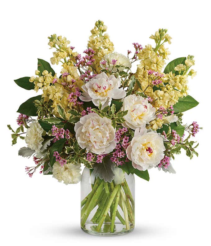 Bouquet of white peonies, soft yellow stock, pink waxflower, and greenery arranged in a clear cylindrical glass vase.