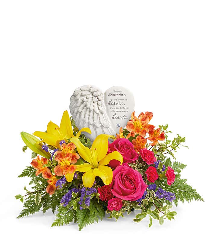 Sympathy flower arrangement with bright yellow lilies, hot pink roses, orange alstroemeria, miniature carnations, and assorted greenery, arranged around a ceramic heart-shaped keepsake with angel wings and an inscription about loved ones in heaven.