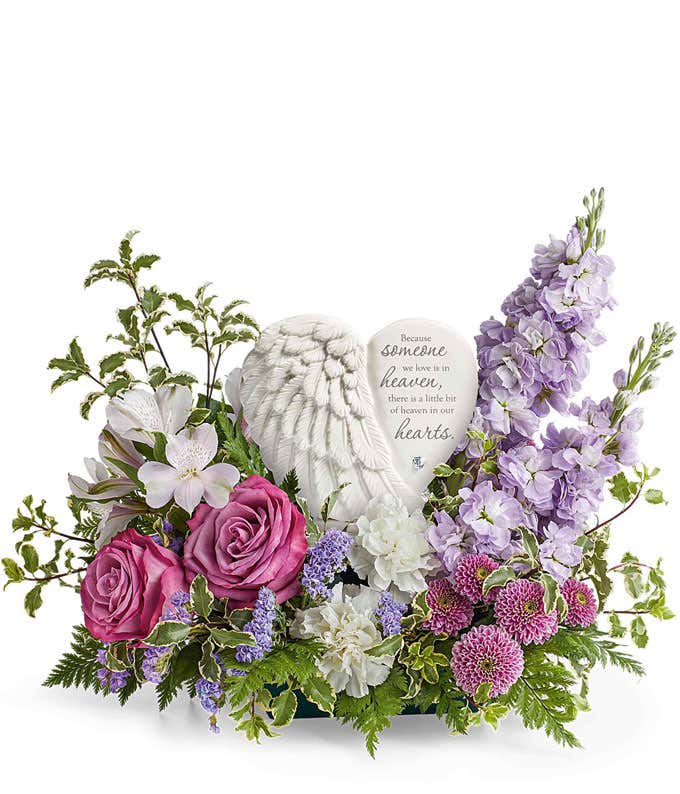 Sympathy flower arrangement with lavender roses, white alstroemeria, white carnations, lavender stock, lavender button spray chrysanthemums, and greenery, surrounding a  heart-shaped keepsake with angel wings and an inscription about loved ones in heaven.