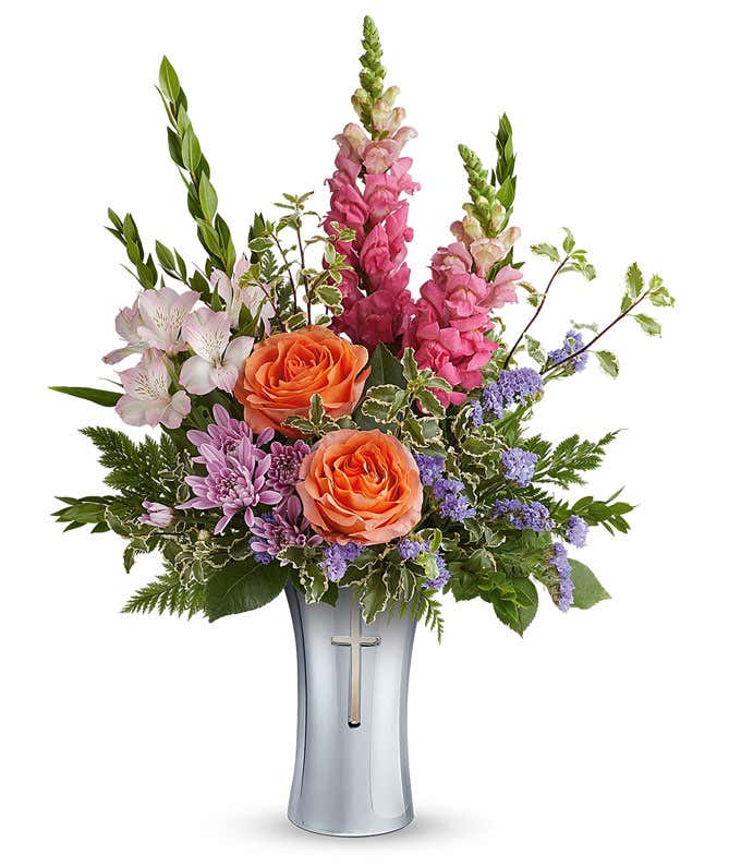 Orange roses, pink snapdragons, pink alstroemeria, lavender cushion spray chrysanthemums, purple statice, and assorted greenery arranged in a keepsake silver cross vase.