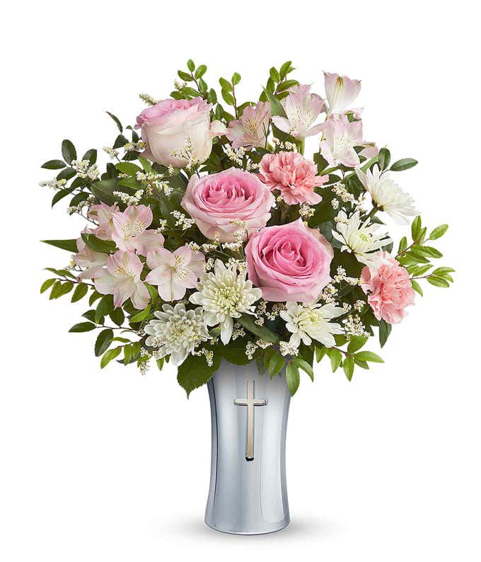 Floral arrangement with pink roses, pink alstroemeria, pink carnations, white cushion spray chrysanthemums, white limonium, and assorted greenery in a keepsake silver vase featuring a cross design.