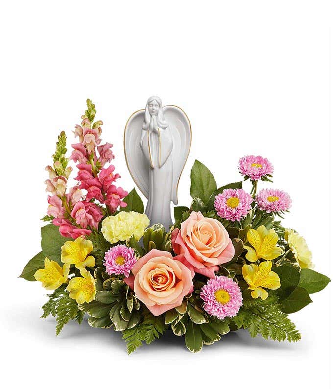 Arrangement featuring peach roses, yellow alstroemeria, yellow carnations, hot pink snapdragons, pink asters, and floral greenery with a ceramic figurine atop the arrangement 