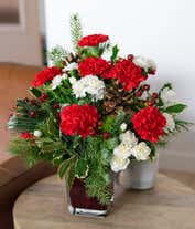Alternative image 4 for A floral arrangement featuring vibrant red carnations and delicate white mini carnations, complemented by lush noble fir and white pine, all elegantly displayed in a red glass cube vase.