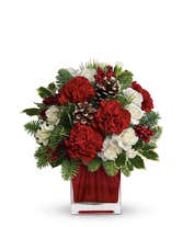 A floral arrangement featuring vibrant red carnations and delicate white mini carnations, complemented by lush noble fir and white pine, all elegantly displayed in a red glass cube vase.