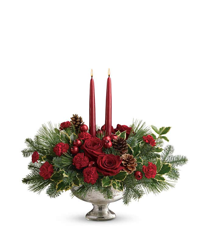 Holiday centerpiece featuring red roses, red miniature carnations, festive berry sprays, pinecones, evergreen greenery, and two red tapered candles arranged in a keepsake mercury glass bowl