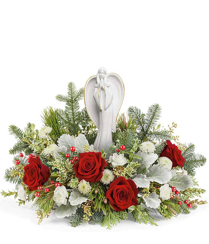 Floral centerpiece featuring red roses, white flowers, silver-hued foliage, and evergreen sprigs, with a serene angel figurine standing in the center.