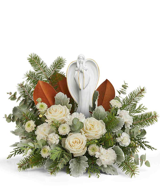 Floral centerpiece featuring white roses, white chrysanthemums, silver-toned foliage, magnolia leaves, and evergreen sprigs, with a peaceful angel figurine at the center.