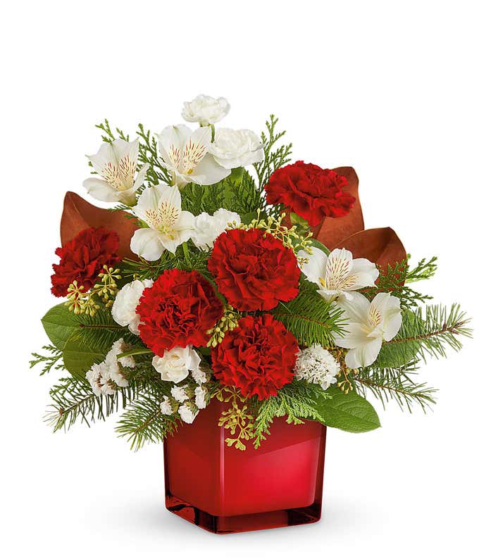 Holiday floral arrangement featuring red roses, red carnations, white alstroemeria, and white miniature carnations, accented with greenery, brown magnolia leaves, and evergreen sprigs, arranged in a red cube vase.