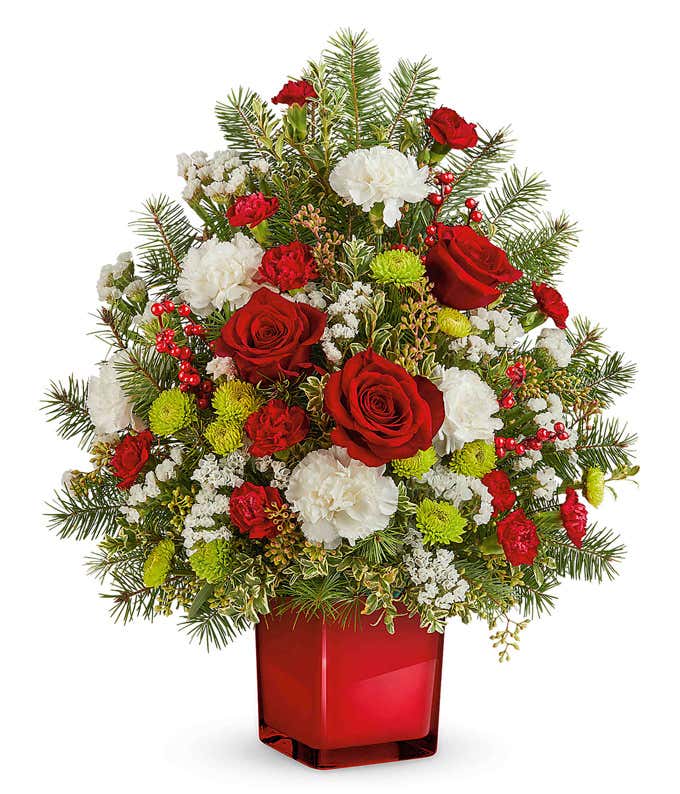  Festive floral arrangement featuring red roses, miniature red carnations, white carnations, green button poms, baby's breath, and seasonal greenery in a red glass cube container. Accented with red berry clusters and pine branches