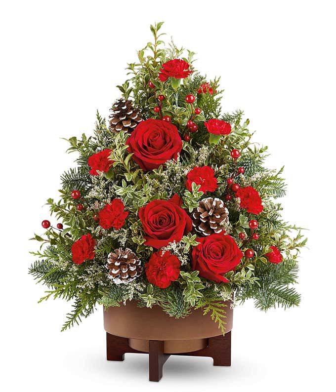Christmas tree-shaped floral arrangement featuring red roses, red carnations, frosted pinecones, red berry accents, and assorted greenery in a brown wooden-style container.