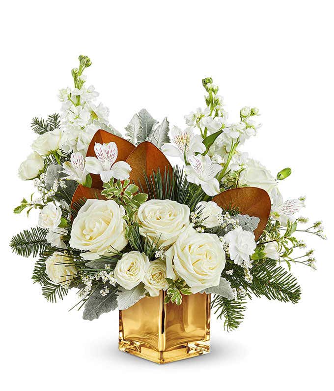 White roses, spray roses, alstroemeria, and stock arranged with evergreens, magnolia leaves, and greenery in a gold cube vase.