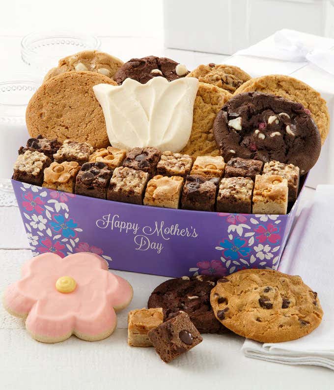 A beautifully arranged gift crate filled with Mrs. Fields cookies and brownie bites, including assorted flavors like triple chocolate, oatmeal raisin, and peanut butter, along with two frosted cookies - a pink daisy and a tulip - adorned with floral desig