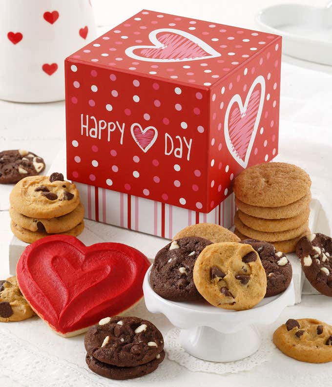 A red gift box with hearts with an assortment of cookies in front with a frosted heart cookie