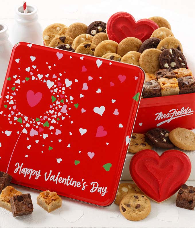 Valentine's Day-themed cookie tin featuring a red container with white and pink heart designs, filled with an assortment of cookies and brownies, including heart-shaped sugar cookies, surrounded by two small bottles of milk.