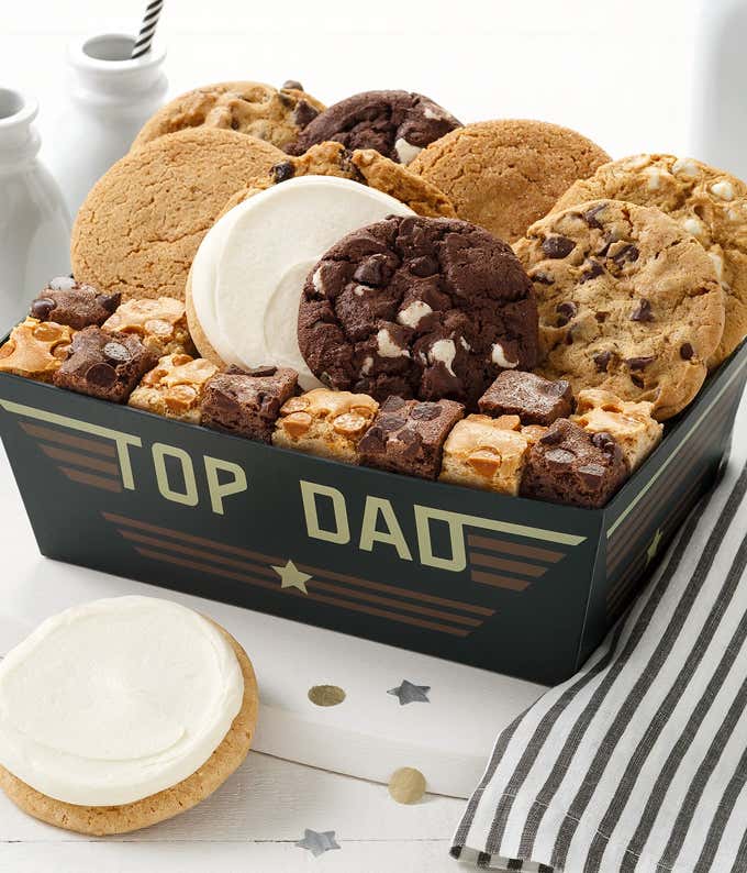 Fathers Day cookie gift box labeled Top Dad, filled with assorted cookies including chocolate chip, oatmeal, sugar with frosting, and chocolate fudge, along with brownie bites, displayed with striped napkin and milk bottles.