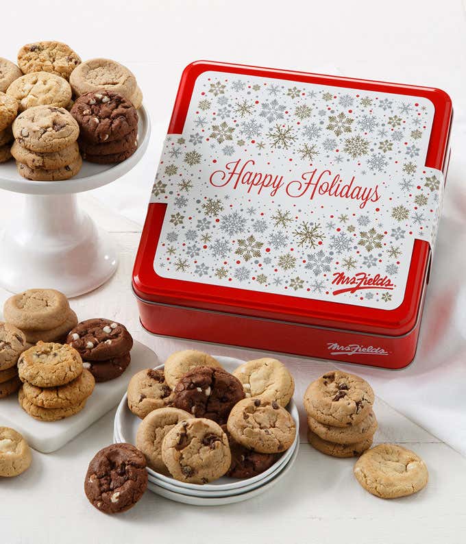 Red holiday cookie tin with a silver snowflake design and “Happy Holidays” written on the lid, surrounded by plates and stands of assorted cookies including chocolate chip, white chocolate macadamia, and triple chocolate varieties.