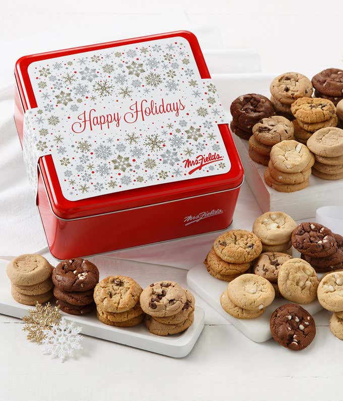 Red holiday cookie tin with a silver snowflake design and “Happy Holidays” message, surrounded by stacks of assorted bite-sized cookies including chocolate chip, triple chocolate, oatmeal raisin, cinnamon sugar, and white chocolate macadamia.