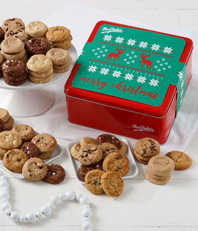 Red holiday cookie tin with a green lid that reads “Merry Christmas” and features white snowflakes and red reindeer, surrounded by plates of assorted bite-sized cookies including chocolate chip, triple chocolate, and white chocolate macadamia.