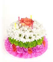 A floral arrangement designed to resemble a birthday cake, featuring layers of white daisies, green button mums, and pink carnations. The top layer is accented with a single peach rose and three striped candles, with a decorative pink lace trim around the