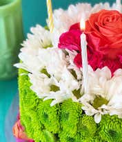 Alternative image 2 for A floral arrangement designed to resemble a birthday cake, featuring layers of white daisies, green button mums, and pink carnations. The top layer is accented with a single peach rose and three striped candles, with a decorative pink lace trim around the