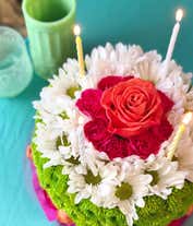 Alternative image 3 for A floral arrangement designed to resemble a birthday cake, featuring layers of white daisies, green button mums, and pink carnations. The top layer is accented with a single peach rose and three striped candles, with a decorative pink lace trim around the