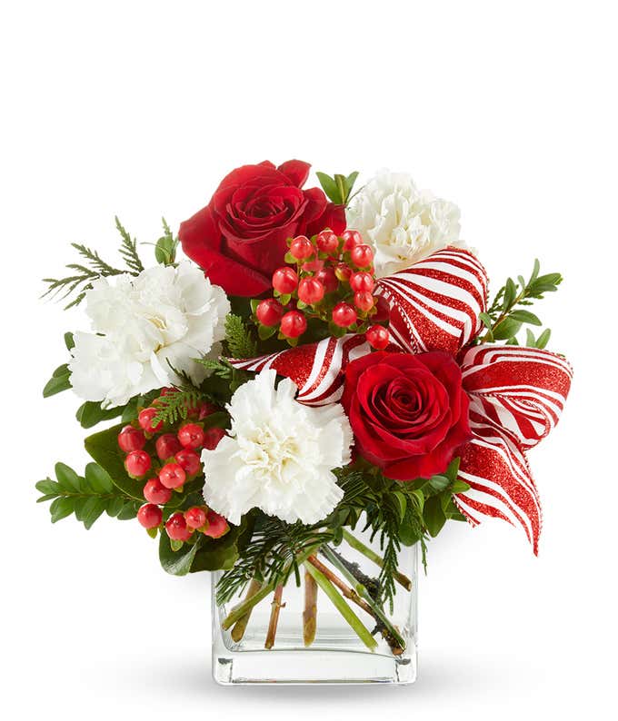 A festive arrangement with red roses, white carnations, red hypericum berries, displayed in a glass cube vase with a decorative ribbon.