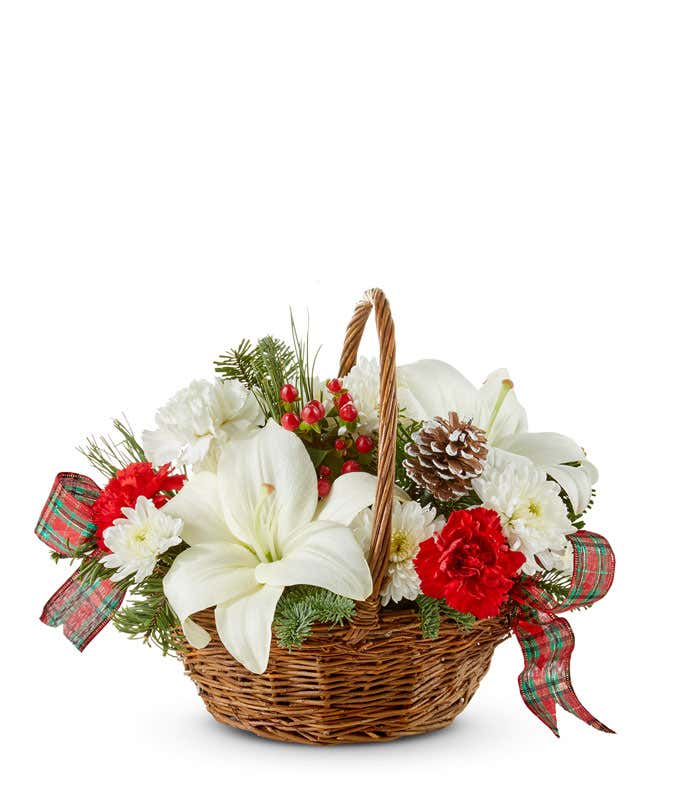 A brown woven basket filled with white lilies, red carnations, red roses, white poms, red berries, and christmas greens with a handle. 