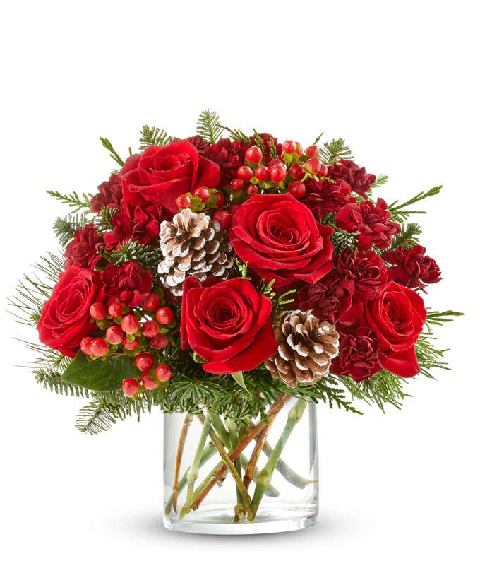 A festive arrangement with red roses, red mini carnations, red hypericum, seasonal greens, displayed in a short glass cylinder vase