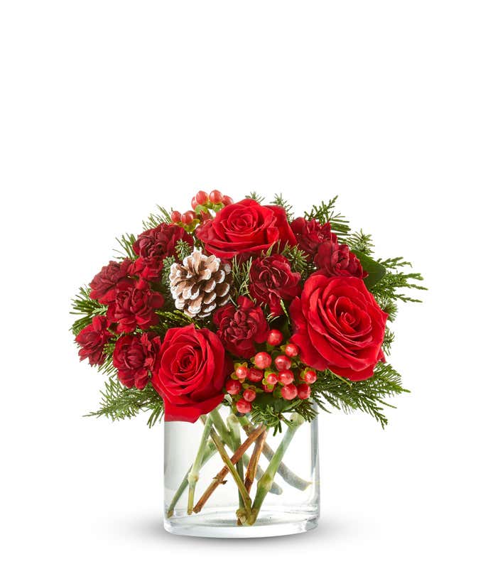 A festive arrangement with red roses, red mini carnations, red hypericum, seasonal greens, displayed in a short glass cylinder vase