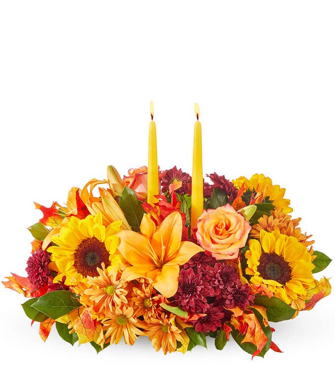 A vibrant autumn centerpiece featuring two tall yellow taper candles surrounded by an arrangement of sunflowers, orange lilies, orange roses, red poms, and assorted greenery.