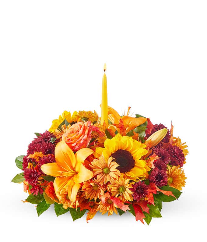 A vibrant autumn centerpiece featuring two tall yellow taper candles surrounded by an arrangement of sunflowers, orange lilies, orange roses, red poms, and assorted greenery.