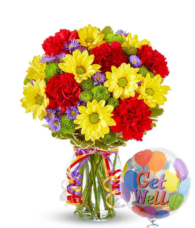 A cheerful floral arrangement featuring red carnations, yellow daisies, purple Monte Casino, and green button poms in a vase decorated with rainbow ribbons, accompanied by a 'Get Well' mylar balloon.