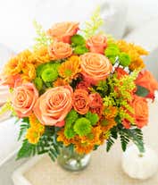 Alternative image 1 for A vibrant floral arrangement featuring orange roses, orange spray roses, butterscotch daisies, green button poms, and solidago, all beautifully displayed in a clear glass vase.