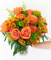 Alternative image 2 for A vibrant floral arrangement featuring orange roses, orange spray roses, butterscotch daisies, green button poms, and solidago, all beautifully displayed in a clear glass vase.