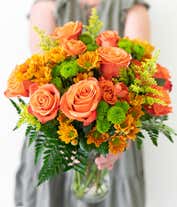 Alternative image 3 for A vibrant floral arrangement featuring orange roses, orange spray roses, butterscotch daisies, green button poms, and solidago, all beautifully displayed in a clear glass vase.