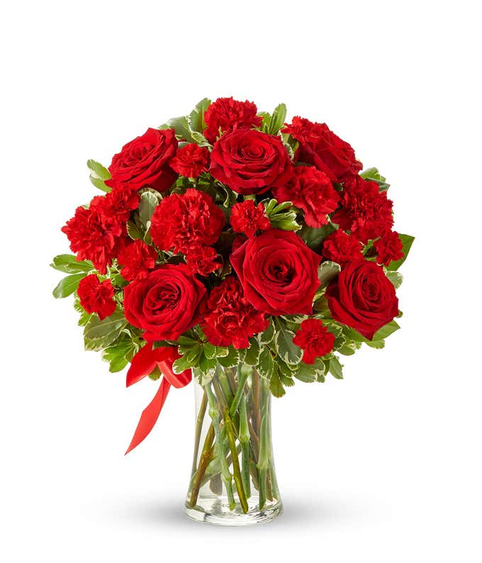 Floral arrangement featuring red roses, red carnations, red mini carnations, and a decorative red ribbon, florist delivered in a clear glass vase.