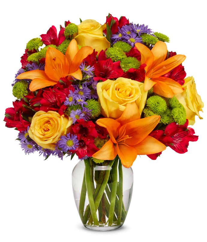 A vibrant bouquet featuring orange lilies, yellow roses, red alstroemeria, purple monte casino, and green button poms, arranged in a clear glass vase.
